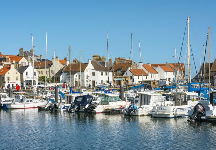 Scenic view of Anstruther Harbour in Fife, Scotland, showing moored fishing boats, calm water, and historic waterfront buildings under a partly cloudy sky.