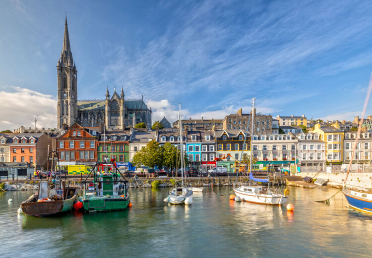 Colorful waterfront buildings and fishing boats in the harbor of Cobh, Ireland, with St. Colman's Cathedral rising in the background under a bright blue sky.
