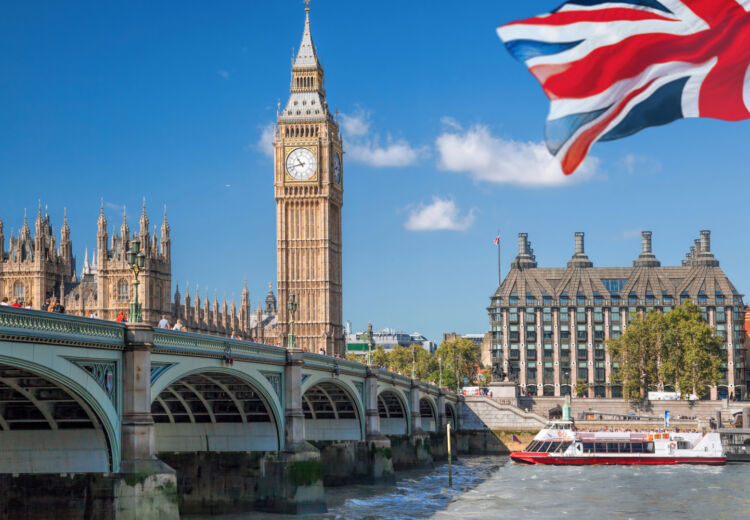 View of Big Ben and the Houses of Parliament in London, with Westminster Bridge spanning the River Thames, a Union Jack flag in the foreground, and a red and white tour boat on the water under a blue sky.