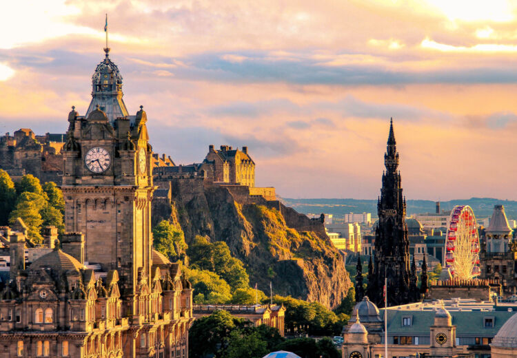 Panoramic view of Edinburgh at sunset, featuring the Balmoral Hotel clock tower, Edinburgh Castle on Castle Rock, the Scott Monument, and a Ferris wheel in the background, with golden light casting a warm glow over the city.
